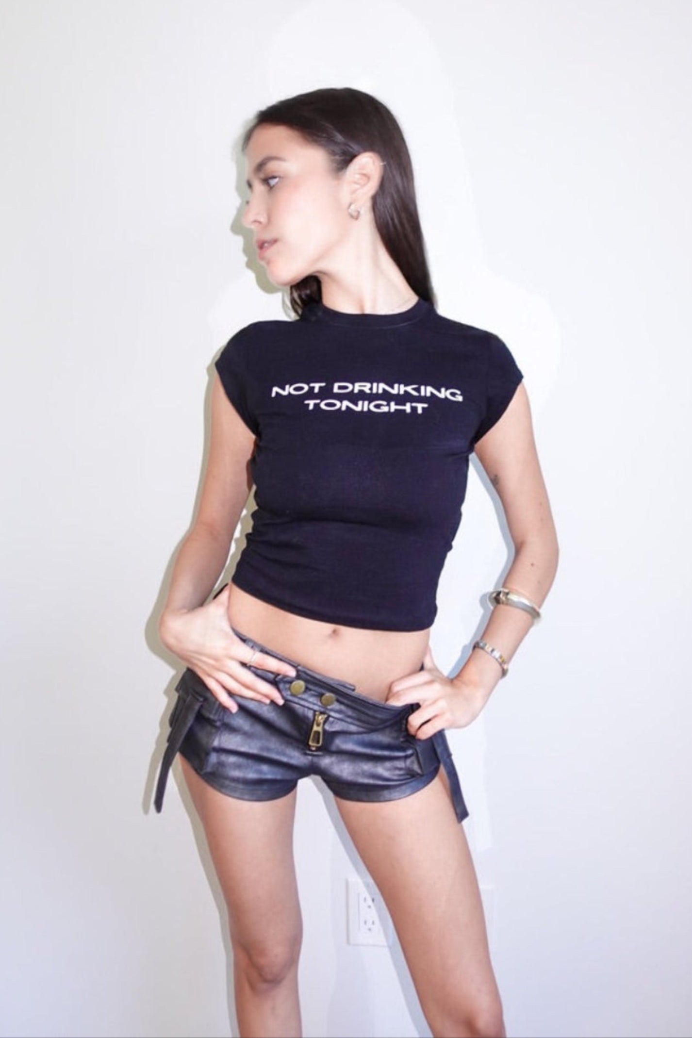 Girl wearing a black croptop slogan babytee with 'Not Drinking Tonight' text and black leather shorts on a white studio background in Miami.